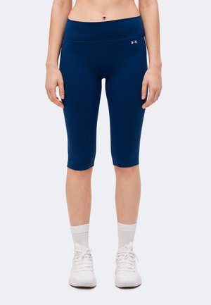 Frau trägt dunkelblaue knielange Under Armour Leggings mit weißen Socken und weißen Sportschuhen, stehend vor einem einfarbigen Hintergrund.