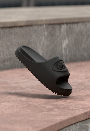 Sort sandal med tekstureret sål og åben tå, placeret på en stenovertag med en sløret grå væg i baggrunden.