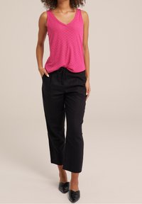 Kvinna iklädd en ärmlös rosa strukturerad V-ringad topp, svarta dragsko-byxor och svarta spetsiga ballerinas, stående med händerna i fickorna.