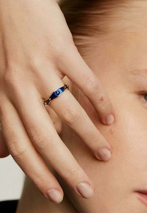 Anillo de oro con un incrustado de esmalte azul, diseño curvado con una textura sutil, adornado con pequeños acentos redondos. Colocado a mano cerca de la cara.
