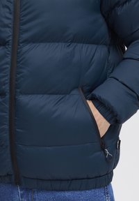 Jūros mėlynos spalvos pūkinė striukė su siuvinėtu raštu, su užtrauktuko kišenėmis, elastingomis rankovėmis ir surištu liemeniu, pagaminta iš lygaus, vandeniui atsparaus audinio.