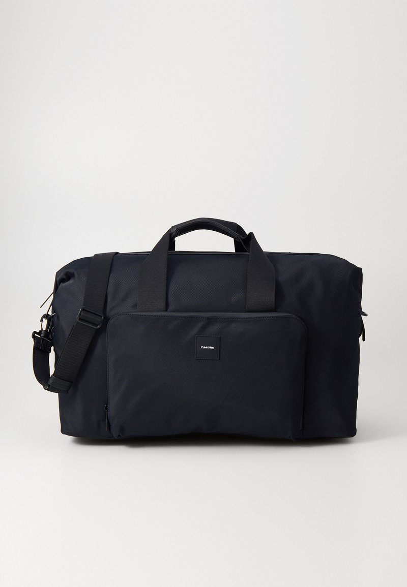 Svart duffelväska i slitstarkt material, med dubbla handtag, en avtagbar axelrem och en framficka med dragkedja och logotyp.