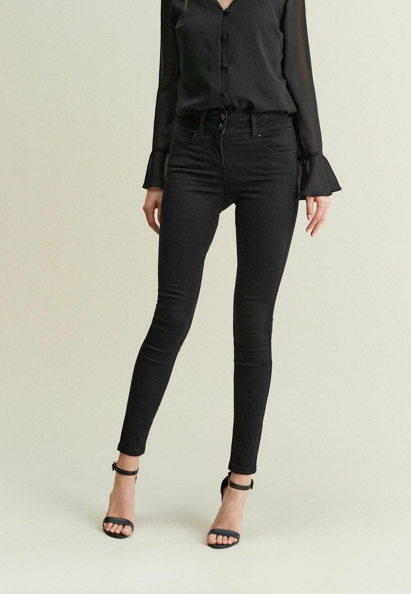 Femme portant un chemisier noir à boutons avec des poignets évasés, un jean skinny noir taille haute et des talons noirs à bride cheville, debout devant un fond uni.