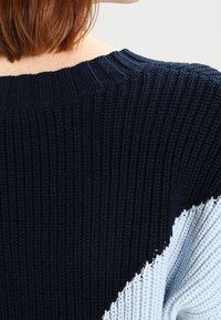 Pletený svetr s kombinací navy modré a světle modré barvy, s žebrovanou strukturou a diagonálním designem s barevnými bloky na rameni.