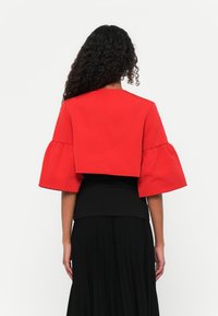 Top rosso corto con ampie maniche a campana, realizzato in un tessuto liscio. Indossato sopra un top nero aderente, abbinato a una gonna nera plissettata.
