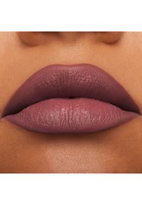 Couleur à lèvres mate dans une teinte mauve avec une finition texturée, présentée sur un fond de peau aux tons chauds. Les lèvres sont pulpeuses avec une forme bien définie.