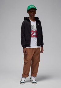 Must musta lukuga tõmblukuga kapuutsiga dressipluus, valge T-särk punase "Jordan 23" kirjaga, pruunid laiad püksid ja roheline baseballi nokamüts "Jordan" tekstiga. Rohelised ja valged spordijalatsid.