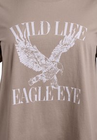 Hellbraunes T-Shirt mit einem weißen Grafikdruck eines Adlers und dem Text "WILD LIFE EAGLE EYE" in einer fetten, abgenutzten Schriftart.