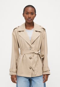 Beige trenchcoat med belte i midjen, dobbelte knapper, brede revers og lange ermer. Stoffet ser glatt ut med en lett glans.