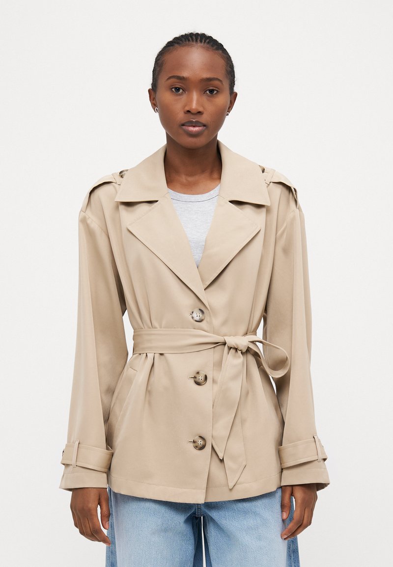 Beige trenchcoat med belte i midjen, dobbelte knapper, brede revers og lange ermer. Stoffet ser glatt ut med en lett glans.