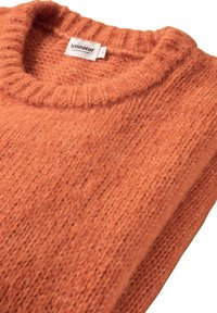Rost-orange gestrickter Pullover mit ribbeligem Rundhalsausschnitt. Der strukturierte Stoff hat lose Fasern, die Wärme und Komfort erhöhen. Etikett sichtbar.