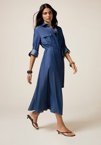 Donna che cammina indossando un vestito midi blu con cintura, maniche arrotolate e sandali neri con tacco, indossando due braccialetti voluminosi.