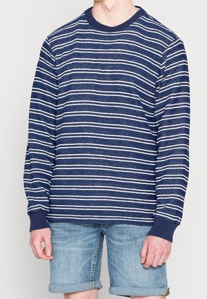 Navy blue long-sleeve sweater with white horizontal stripes, ribbed neckline, and cuffs; paired with light denim shorts.