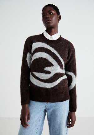 Bruine gebreide trui met een wit abstract patroon, met een geribde halslijn en mouwen, gedragen over een witte colbert.
