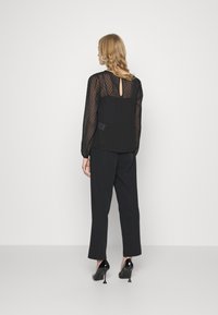 Blusa preta transparente com padrão de bolinhas, detalhe de abertura nas costas, usada com calças largas pretas e sapatos de salto alto pretos.