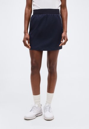 Žena v tmavomodrej športovej sukni, bielych ponožkách a bielych teniskách, stojaca na svetlom jednoduchom pozadí.