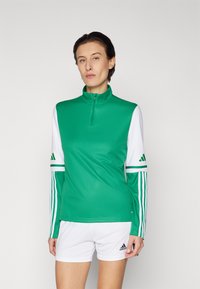 Grøn zip-up træningstrøje med hvide ærmer, der har tre sorte striber på hver arm. Materialet er glat og tætsiddende. Bæres med hvide shorts.
