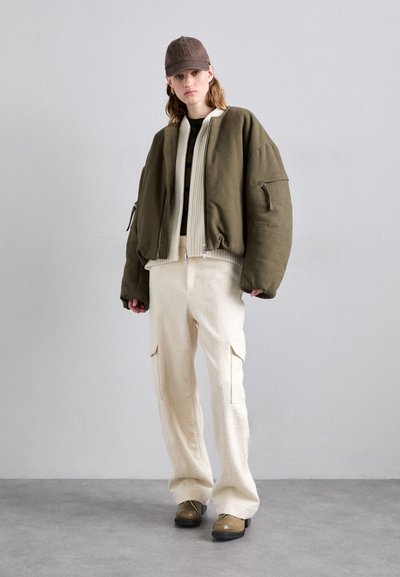 Giacca bomber oversize verde oliva con chiusura con zip, inserti in rilievo beige, abbinata a pantaloni testurizzati crema e berretto marrone.