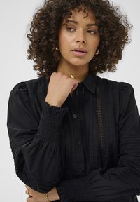 Femme aux cheveux bouclés portant un blouse noire avec des détails en dentelle, tenant sa main près de son menton, portant une bague en or et des boucles d'oreilles.