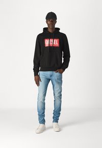 Black hoodie with a red "DIESEL" logo, paired with light blue jeans and white sneakers. Model wears a black cap. Plain background.