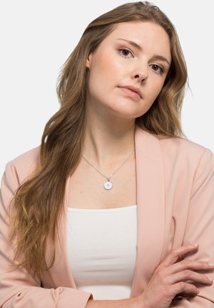 Silberne Halskette mit einem runden Anhänger, der ein strukturiertes Design aufweist, getragen über einem weißen Oberteil. Das Model hat lange Haare und trägt ein pinkes Blazer.