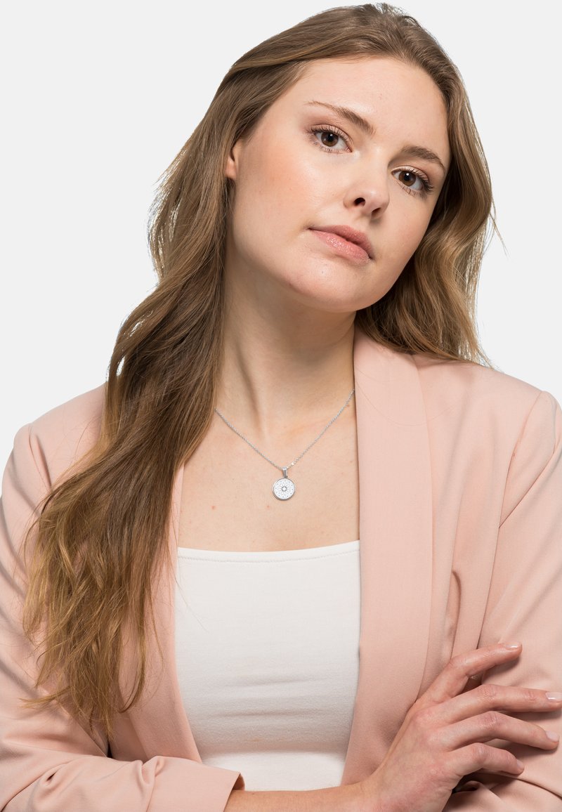 Collana d'argento con un pendente circolare caratterizzato da un design testurizzato, indossata su un top bianco. Il modello ha capelli lunghi ed è vestito con un blazer rosa.