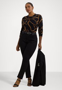 Maglione nero con motivo a catena dorata, jeans neri con una cintura e sandali neri. La modella tiene un blazer nero.