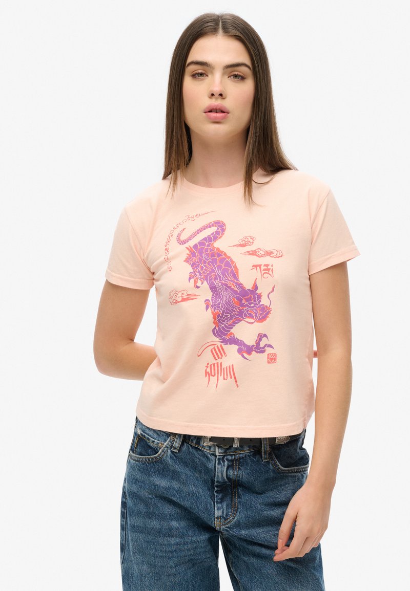 Camiseta corta de algodón melocotón con un gráfico de dragón púrpura y rojo, con mangas cortas y un cuello redondo, combinada con jeans de mezclilla azul.