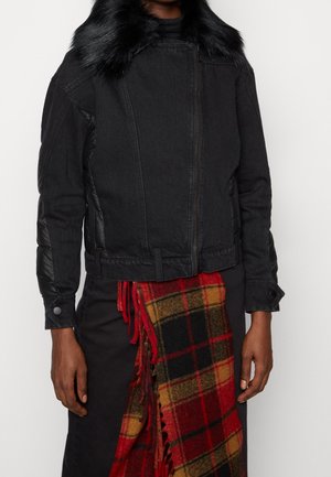 Personne portant une veste noire avec col en fourrure et pantalon noir, tenant une écharpe à carreaux rouge et jaune avec des franges.