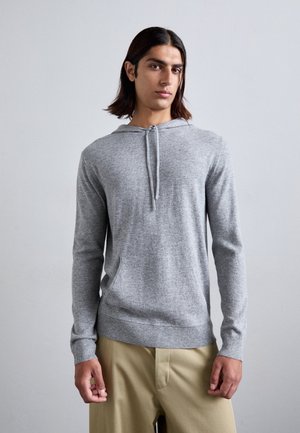 Sweatshirt à capuche gris avec cordon de serrage, manches longues, poche avant et ourlet côtelé. Porté par-dessus un pantalon clair.