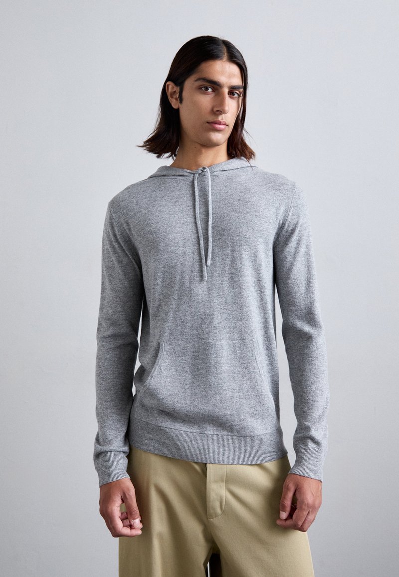 Grauer Kapuzenpullover mit Kordelzug, langen Ärmeln, aufgesetzter Tasche und geripptem Saum. Über hellen Hosen getragen.