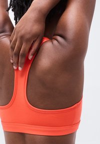 Personne à la peau foncée portant un soutien-gorge de sport orange, étirant le bras derrière la tête sur un fond clair uni.