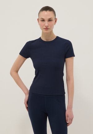 Frau steht mit einer Hand in der Hüfte, trägt ein tailliertes, marineblaues Kurzarmhemd und passende Hose vor einem schlichten hellen Hintergrund.