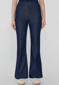 Pantalones vaqueros de campana de color azul oscuro, fabricados en denim, con cintura alta, cierre de botón en la parte delantera, dos bolsillos laterales y un dobladillo ligeramente desgastado.