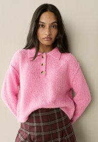 Šviesiai rožinė puri džemperis su apykakle, trimis mygtukais ir laisvu kirpimu. Derinama su tamsiai langiniu sijonu, turinčiu raukinukų.