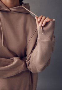 Brązowa bluza z kapturem wykonana z miękkiego materiału, z kapturem ze sznurkiem i ściągaczami z otworami na kciuki. Zbliżenie na rękaw i dłoń.