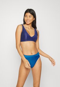 Námořnické modré bikiny s topem ve tvaru V s vlnitým vzorem; odpovídající vysoké kalhotky v jednobarevné modré. Hladká textura, minimální design.
