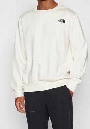 Persona che indossa una felpa bianca a maniche lunghe con il logo The North Face sul petto e pantaloni neri, mano in tasca.