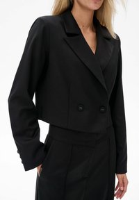 Czarna skrojona żakietka z podwójnym zapięciem, klapami wciętymi i dwoma czarnymi guzikami. Gładka tekstura, dopasowany krój, usztywniane rękawy.