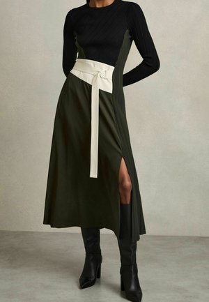 Femme portant une robe noire côtelée à manches longues avec jupe vert foncé, ceinture blanche, fente latérale et bottes noires montantes à talons.