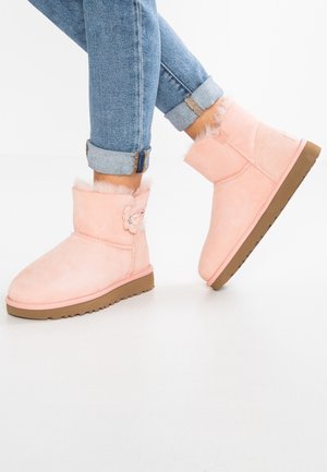 Legs in rolled-up blue jeans wearing pink fur-lined ankle boots with floral embellishment and brown rubber soles on white floor.