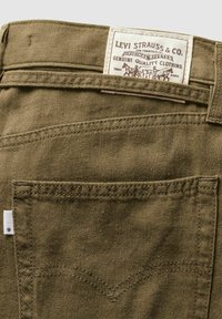 Olivengrønne bukser med tekstureret stof, der har et hvidt Levi Strauss & Co. mærke og dobbeltstikninger på baglommens dekor.
