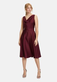 Bordeaux satijnen jurk met een coul neckline en A-lijn silhouet. De details omvatten een aansluitende taille en een vloeiende rok, gecombineerd met zilveren hakken.