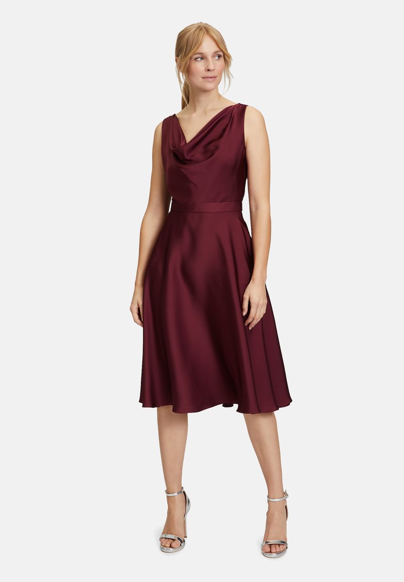 Bordeaux satijnen jurk met een coul neckline en A-lijn silhouet. De details omvatten een aansluitende taille en een vloeiende rok, gecombineerd met zilveren hakken.