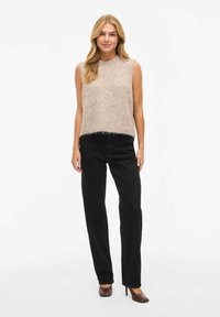 Bēša pūkaina krekla bez piedurknēm ar apaļu izgriezumu, kombinēts ar melnām taisnstūrveida biksēm un brūnām augstpapēžu kurpēm.