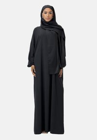 Schwarze, langärmelige Abaya in voller Länge mit fließendem Schnitt. Glatter Stoff, schichtweise Design und passender Hijab. Kombiniert mit minimalen Accessoires.