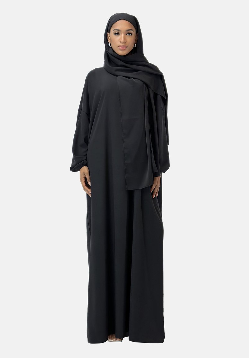 Schwarze, langärmelige Abaya in voller Länge mit fließendem Schnitt. Glatter Stoff, schichtweise Design und passender Hijab. Kombiniert mit minimalen Accessoires.
