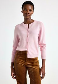 Lyserød cardigan i blødt strik, rund hals, lukning med frontknapper, ribbede manchetter, parret med brune fløjlbukser. Glat tekstur, tætsiddende design.
