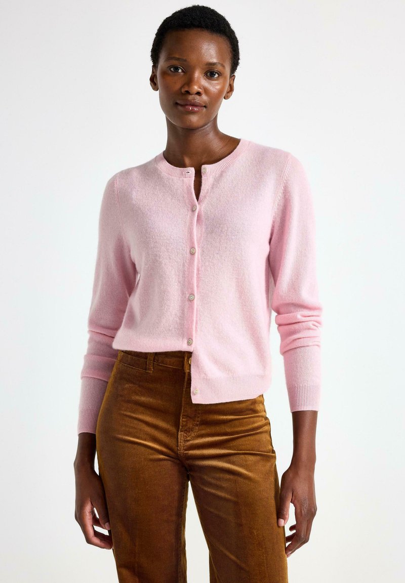 Lyserød cardigan i blødt strik, rund hals, lukning med frontknapper, ribbede manchetter, parret med brune fløjlbukser. Glat tekstur, tætsiddende design.