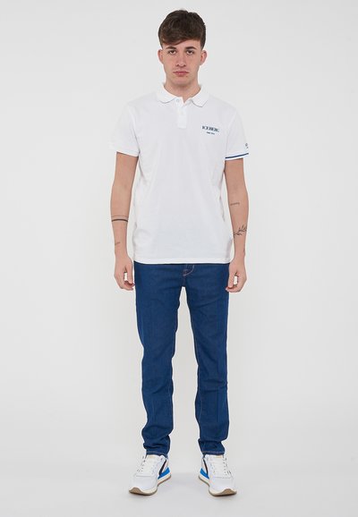Polo bianco con logo blu, maniche corte con dettagli a righe blu e bianche, abbinato a jeans blu scuro e sneakers bianche.